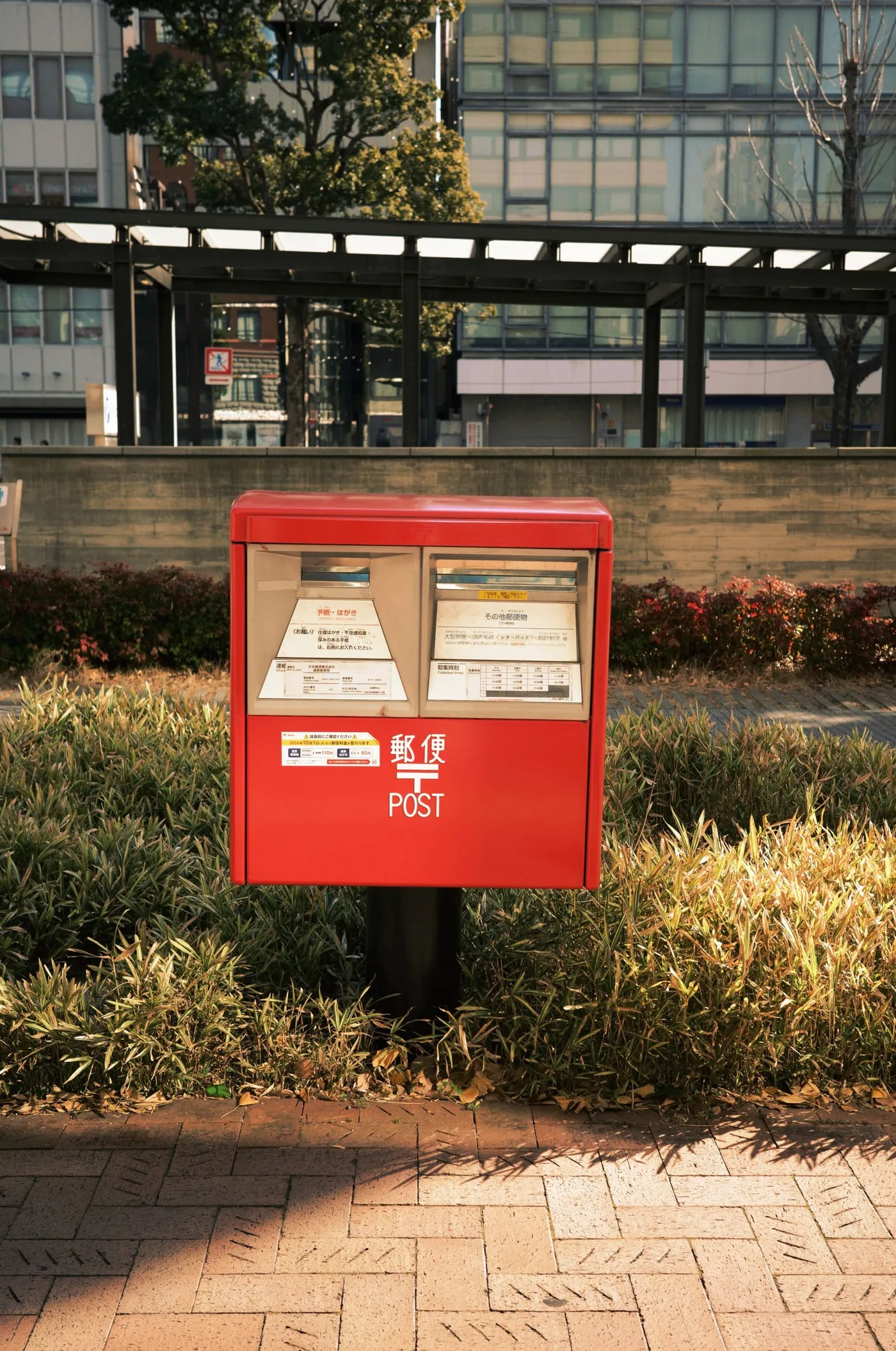 A red Japanese post box on a paved street corner, with printed collection-time notices posted on its front.