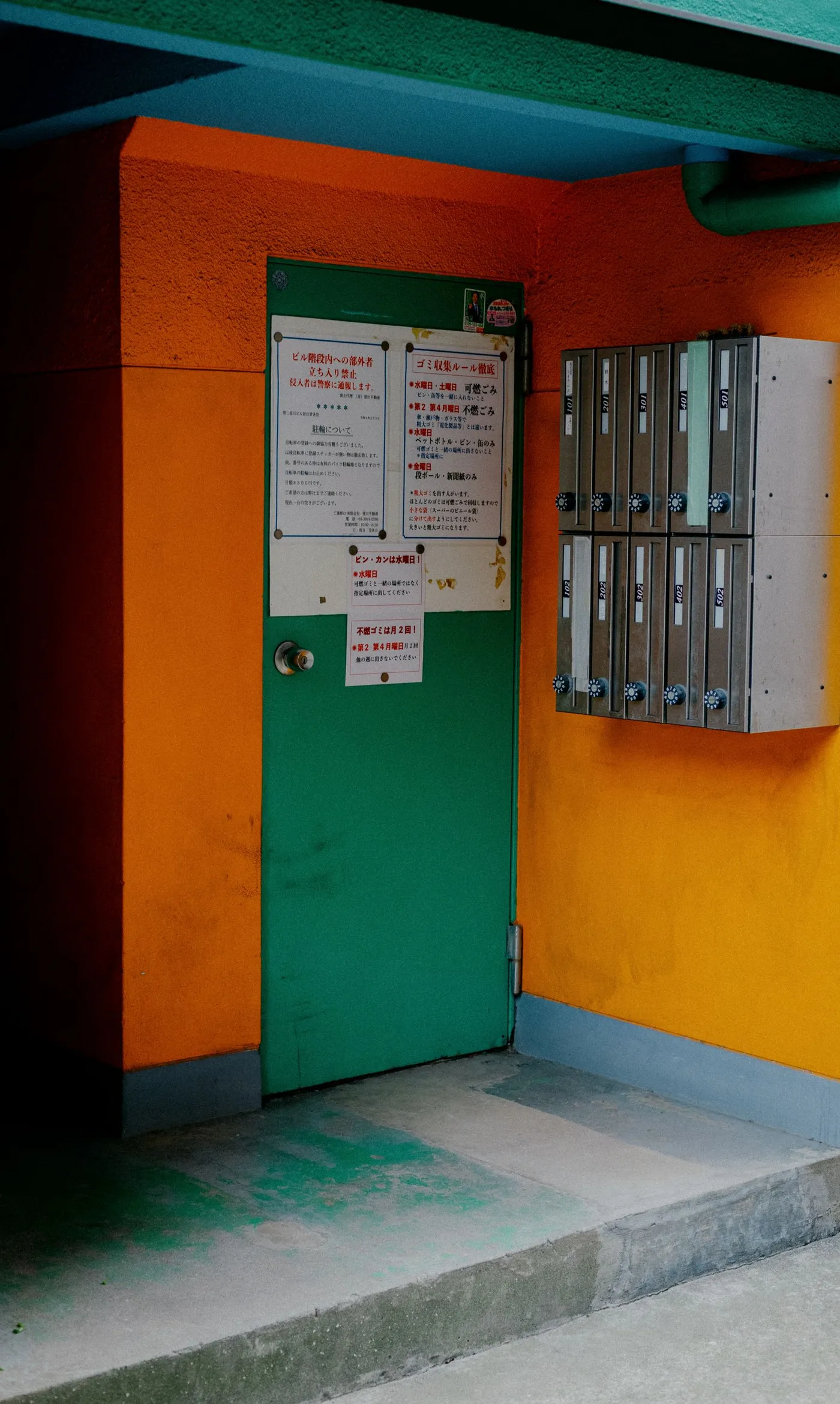Apartment mailboxes mounted on an orange wall beside a green door with printed notices, under a covered entryway.