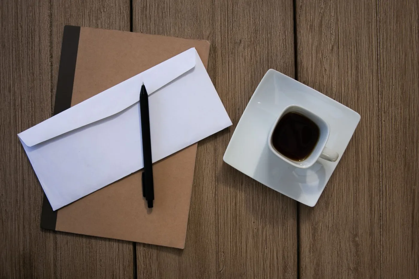 A white envelope and a pen resting on a kraft-colored notebook beside a small cup of coffee on its saucer.