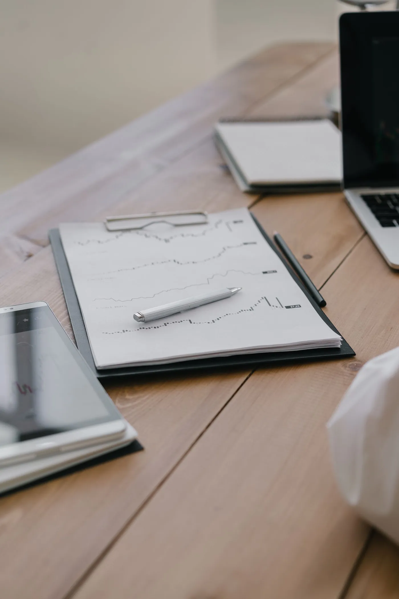 A wooden desk with a clipboard holding a hand-drawn line chart, a pen and a pencil beside it, a spiral notebook and the edge of an open laptop in the background, and a tablet-sized device in the foreground.