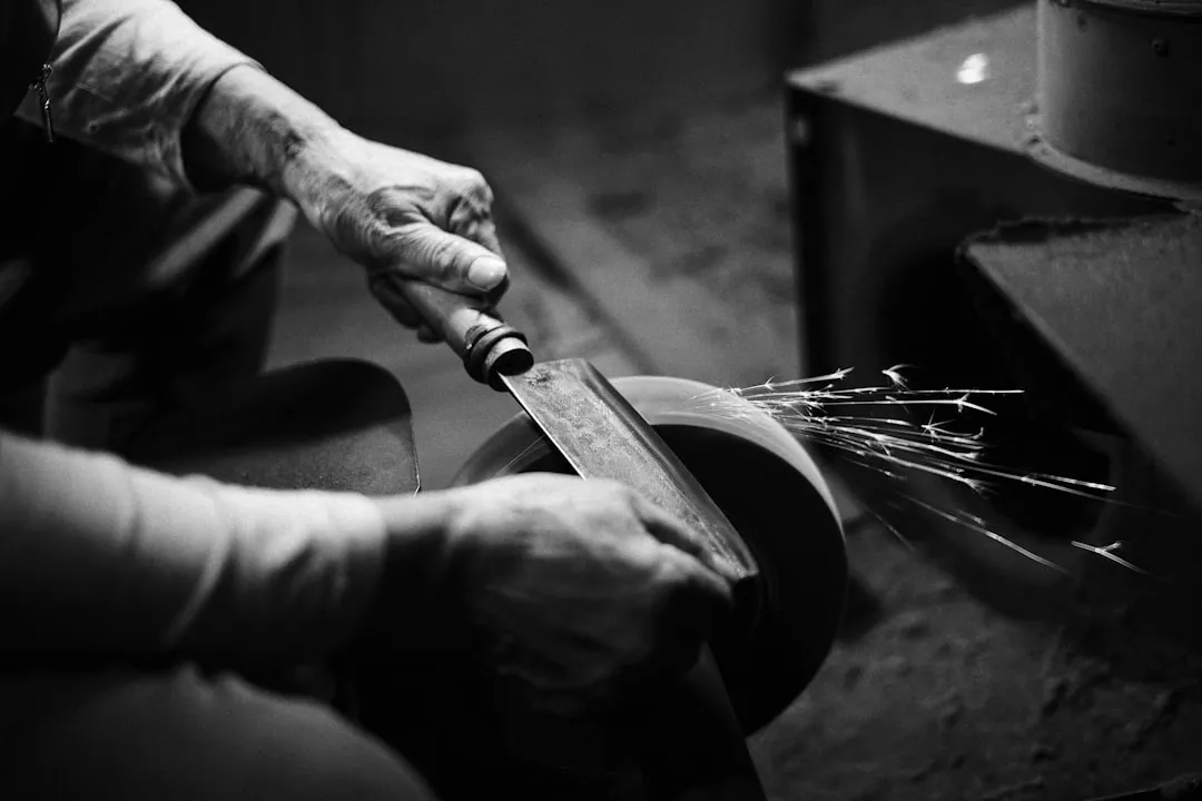 A worker holding a wide blade against a powered grinding wheel in a workshop.