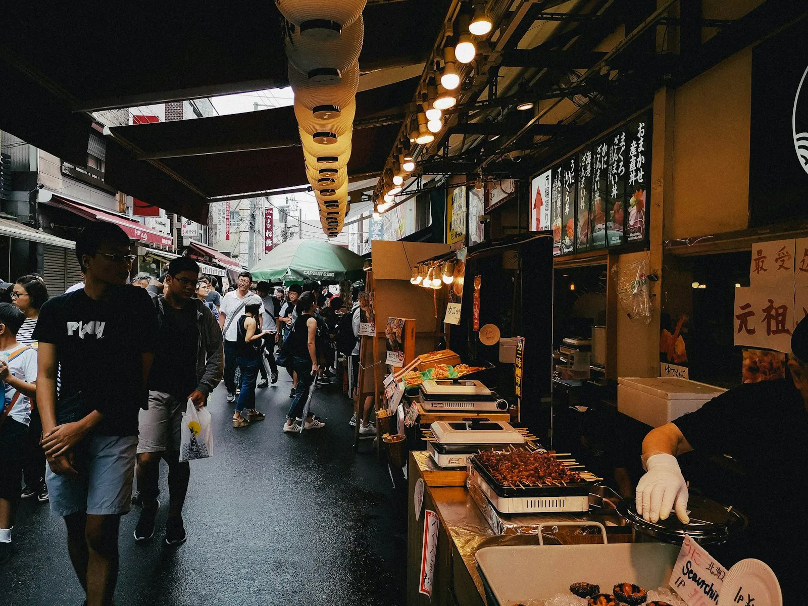 Shoppers walking past food stalls in a covered market street.