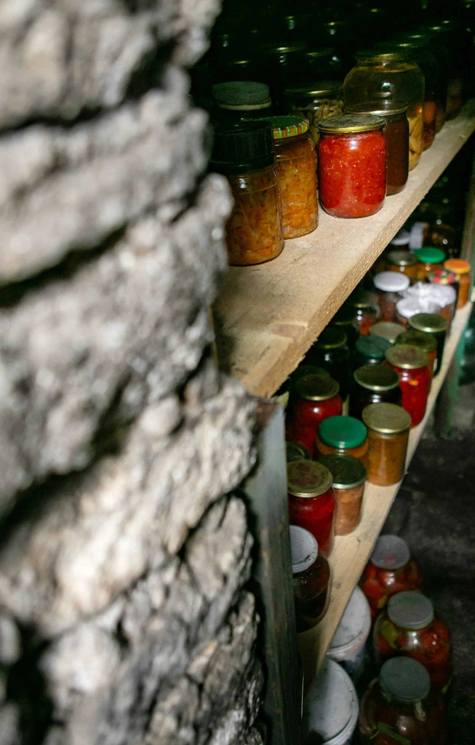 Glass jars of preserved food lined up on wooden shelves inside a stone-walled storage room.