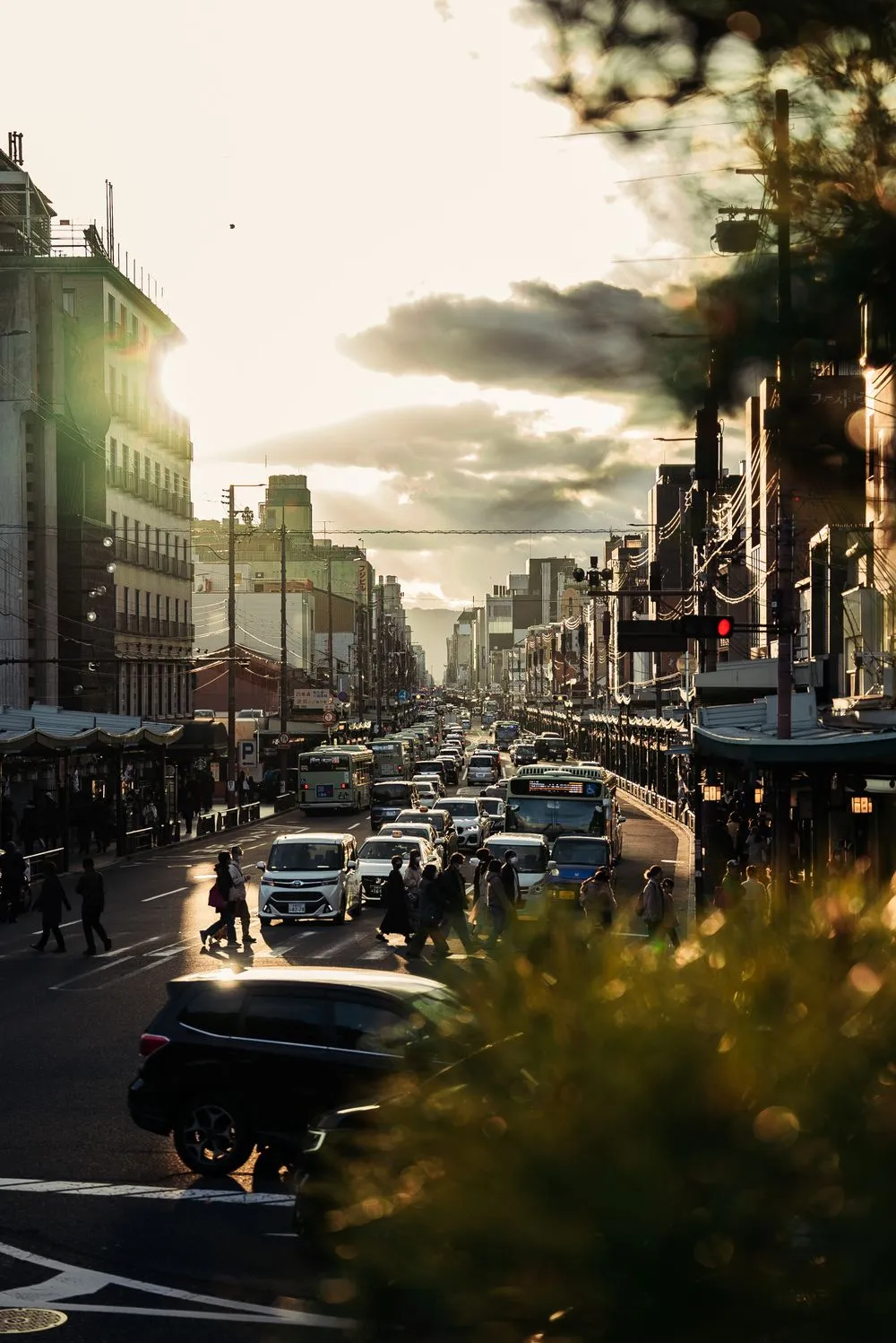 A busy street in Kyoto at dusk with cars, buses and people crossing at a signal.