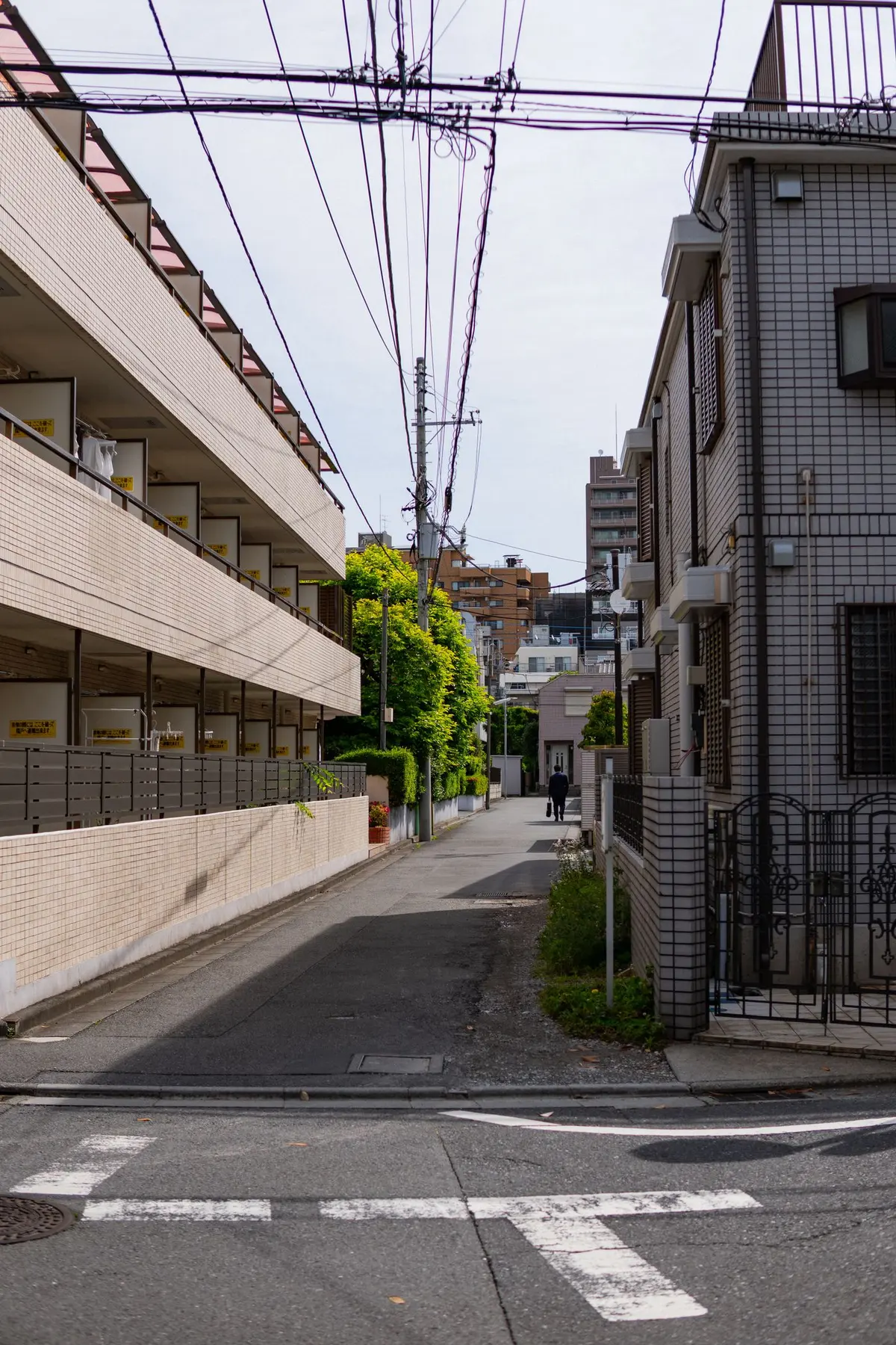 A quiet residential street in Japan with low apartment buildings, overhead power lines and a person walking in the distance.