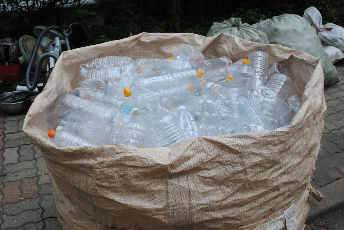 A large woven sack filled with rinsed clear plastic bottles, with more filled sacks behind it on a paved surface.