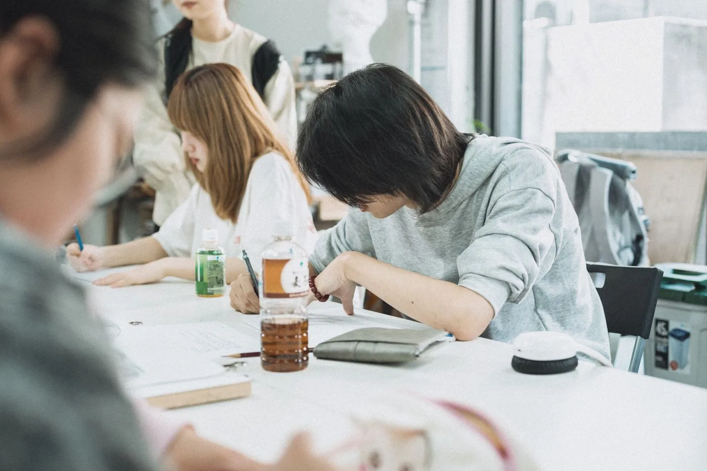 Three people sitting along a table in a bright room, writing in notebooks with drinks and pencil cases in front of them.