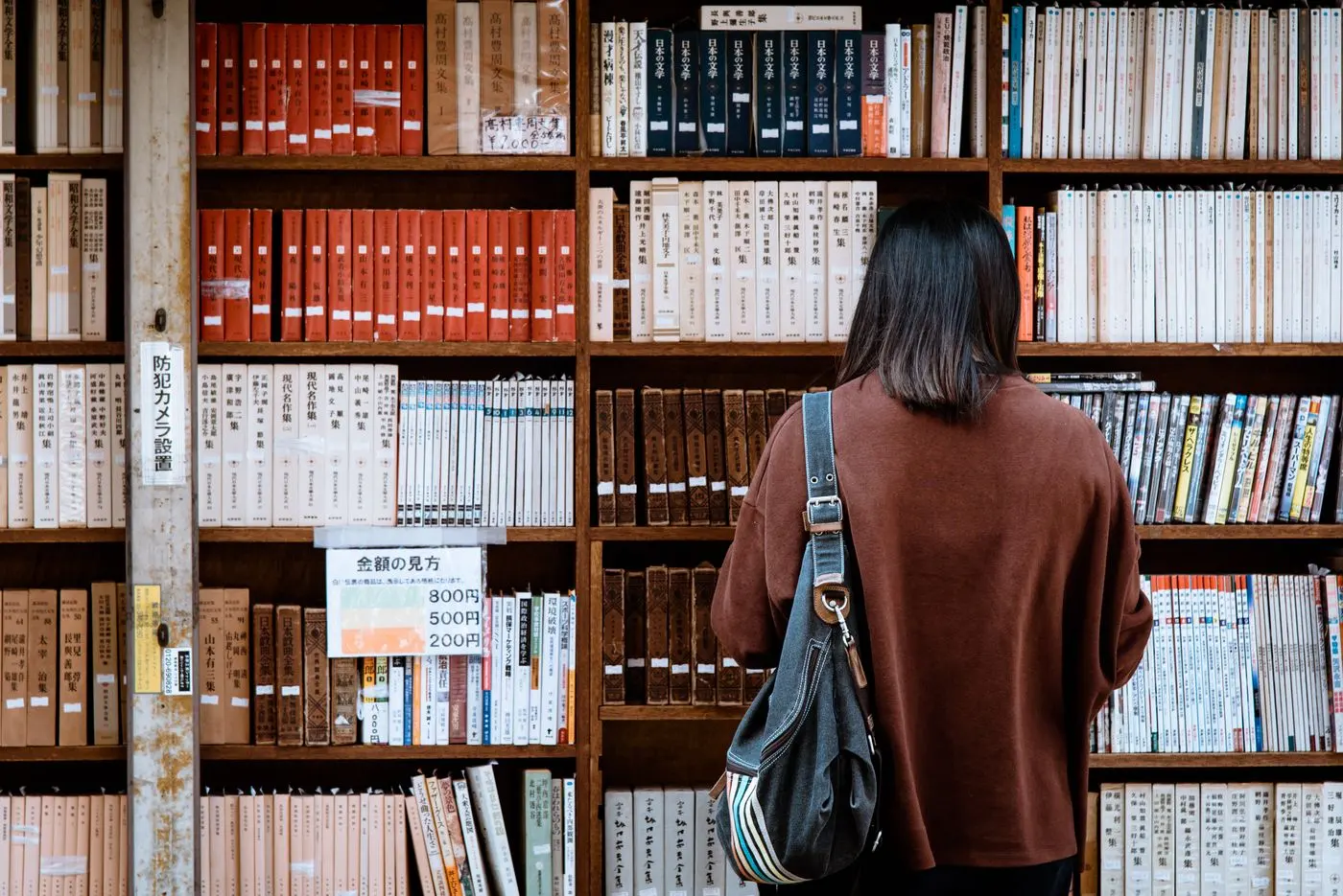 A wall of shelves in a Japanese second-hand bookshop, packed with paperbacks, with a person standing in front of it seen from behind.