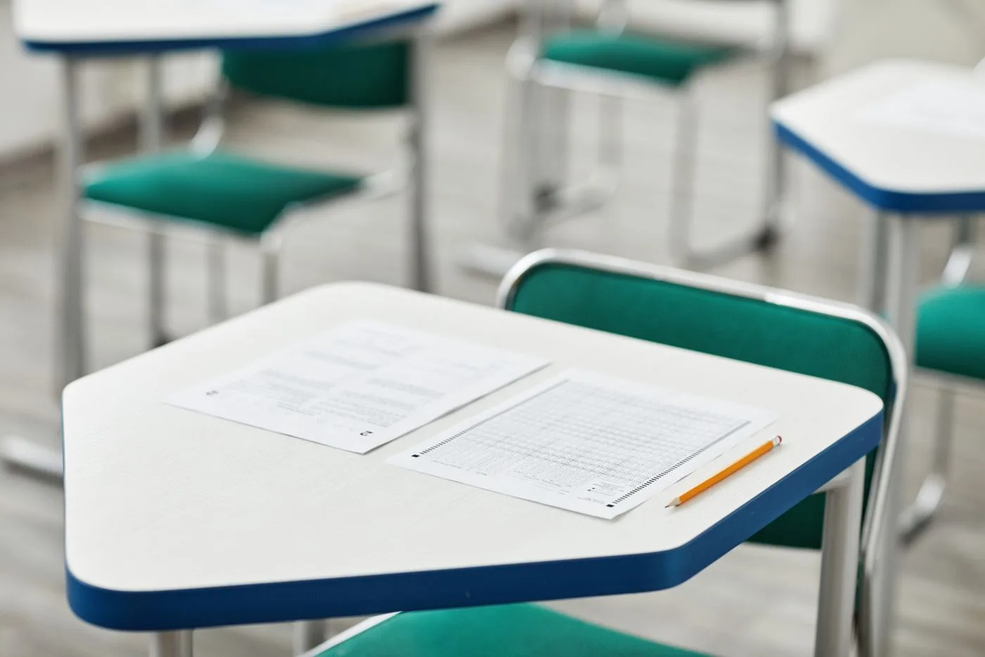 An empty classroom desk with an exam answer sheet, a question paper and a sharpened pencil laid out on it.
