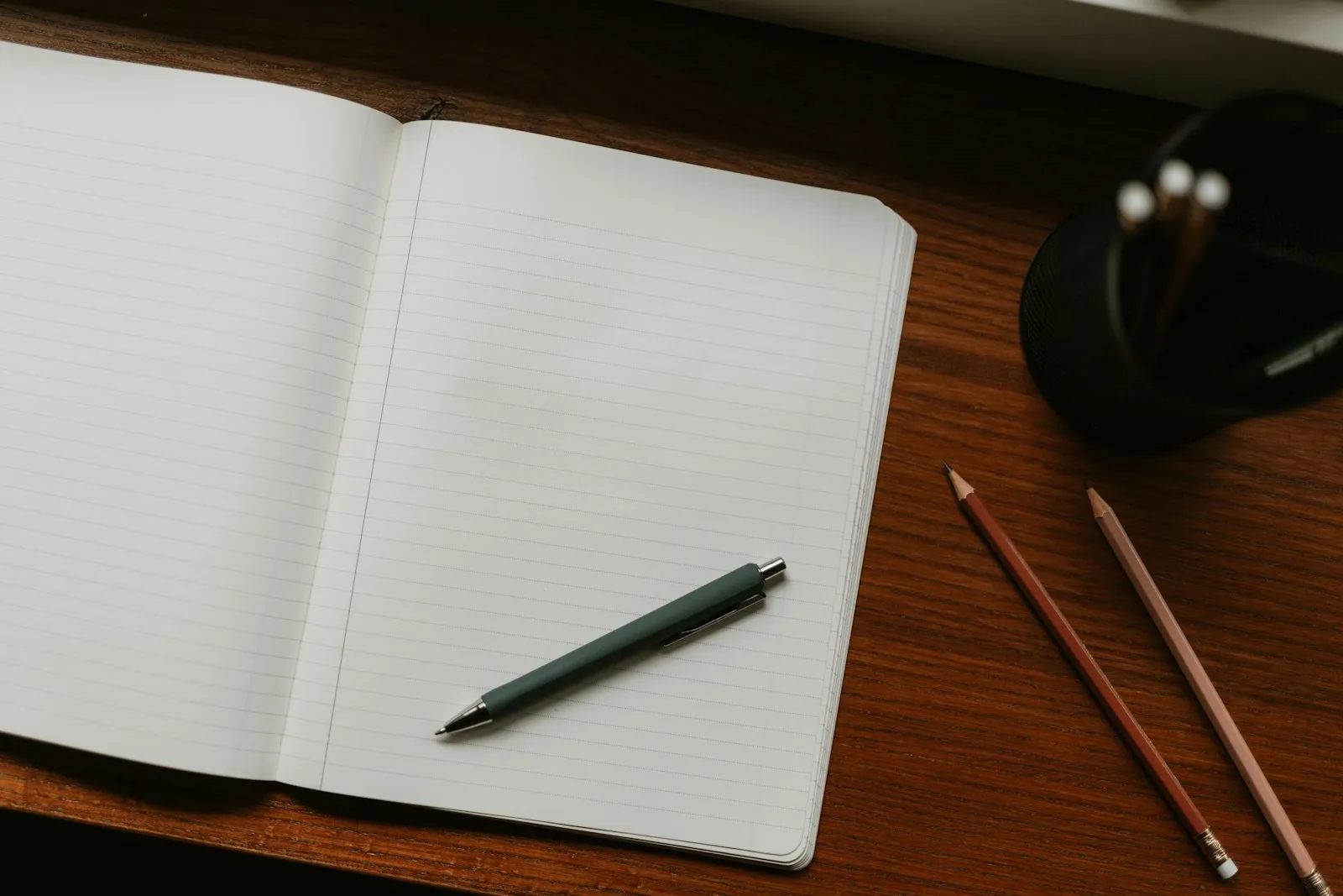 An open notebook with a pen and pencils on a desk.