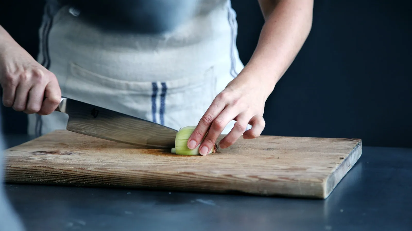 A cook slicing a green vegetable on a wooden chopping board with a kitchen knife.