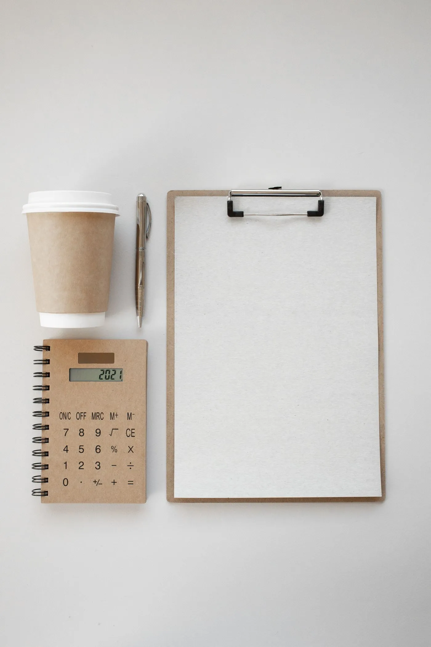 A flat lay of a blank clipboard, a paper cup of coffee, a pen, and a small calculator on a white surface.