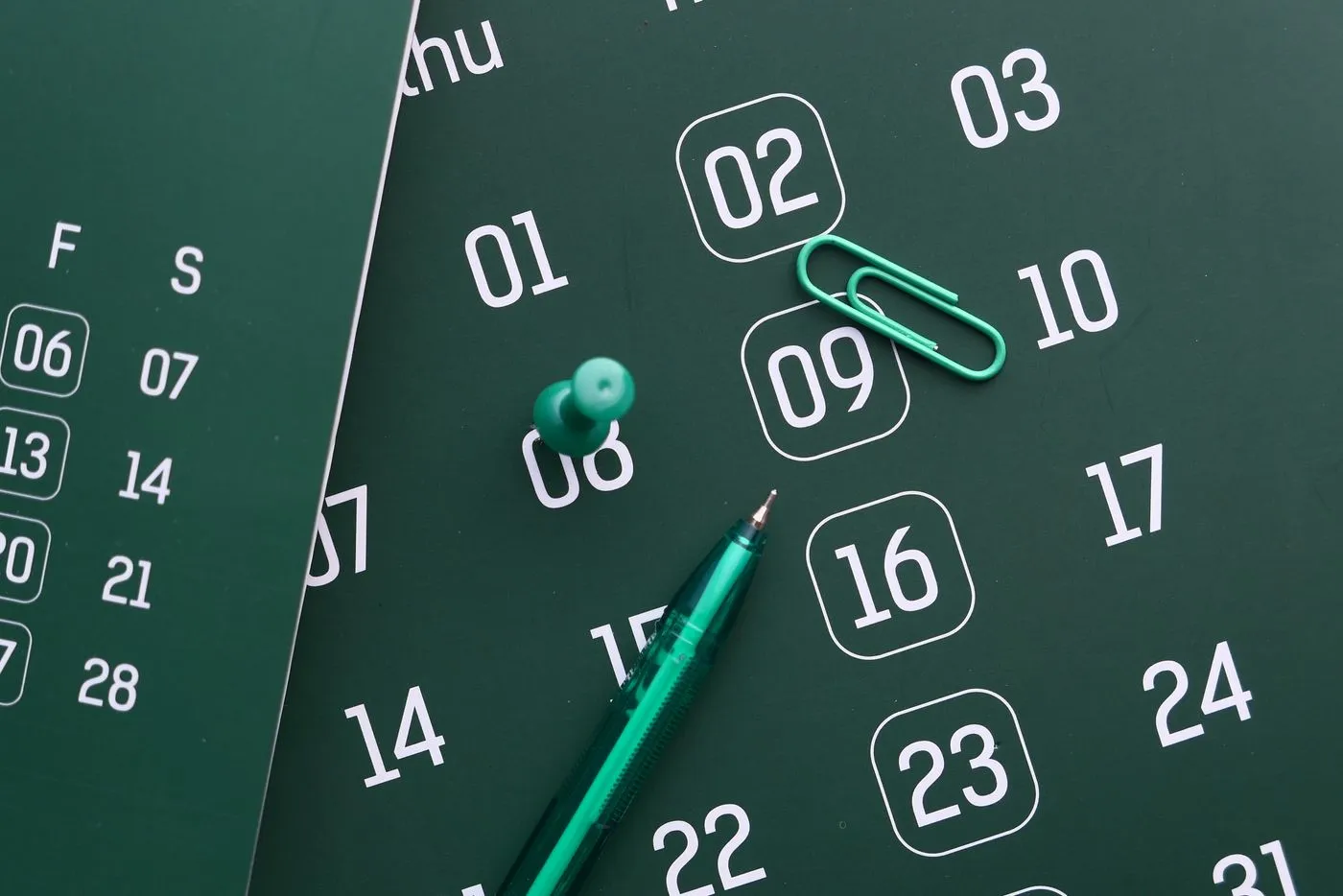 A dark green calendar page showing numbered days, with a green pushpin, paperclip and pen resting on it.