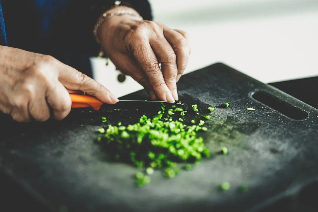 Hands chopping spring onions into small pieces on a dark cutting board.