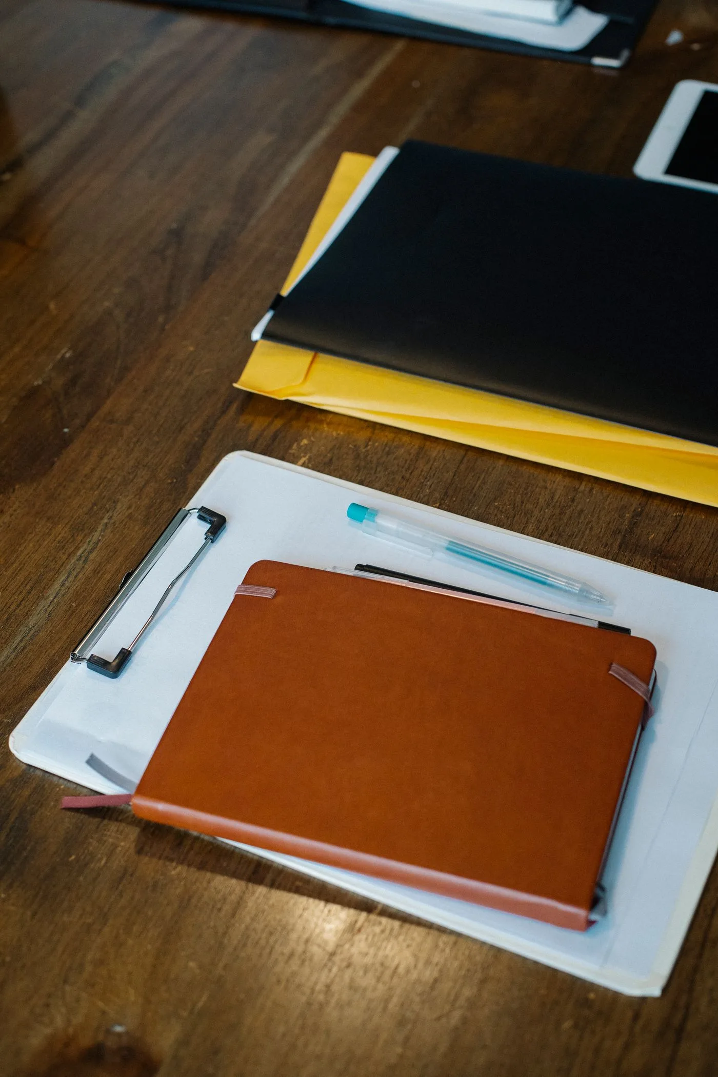 A leather-covered notebook resting on a clipboard of blank paper, with two pens, a black folder and a yellow envelope on a dark wooden table.