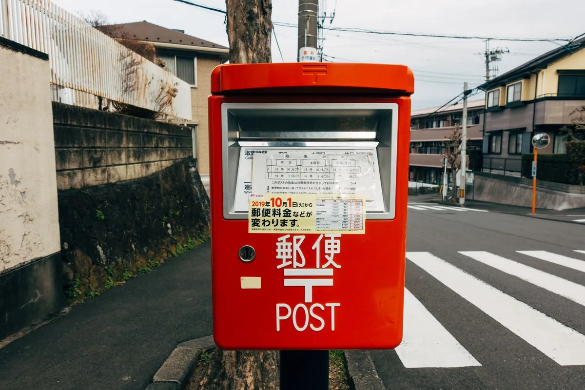 A red Japanese post box on a residential street corner, with a collection timetable posted on its front.
