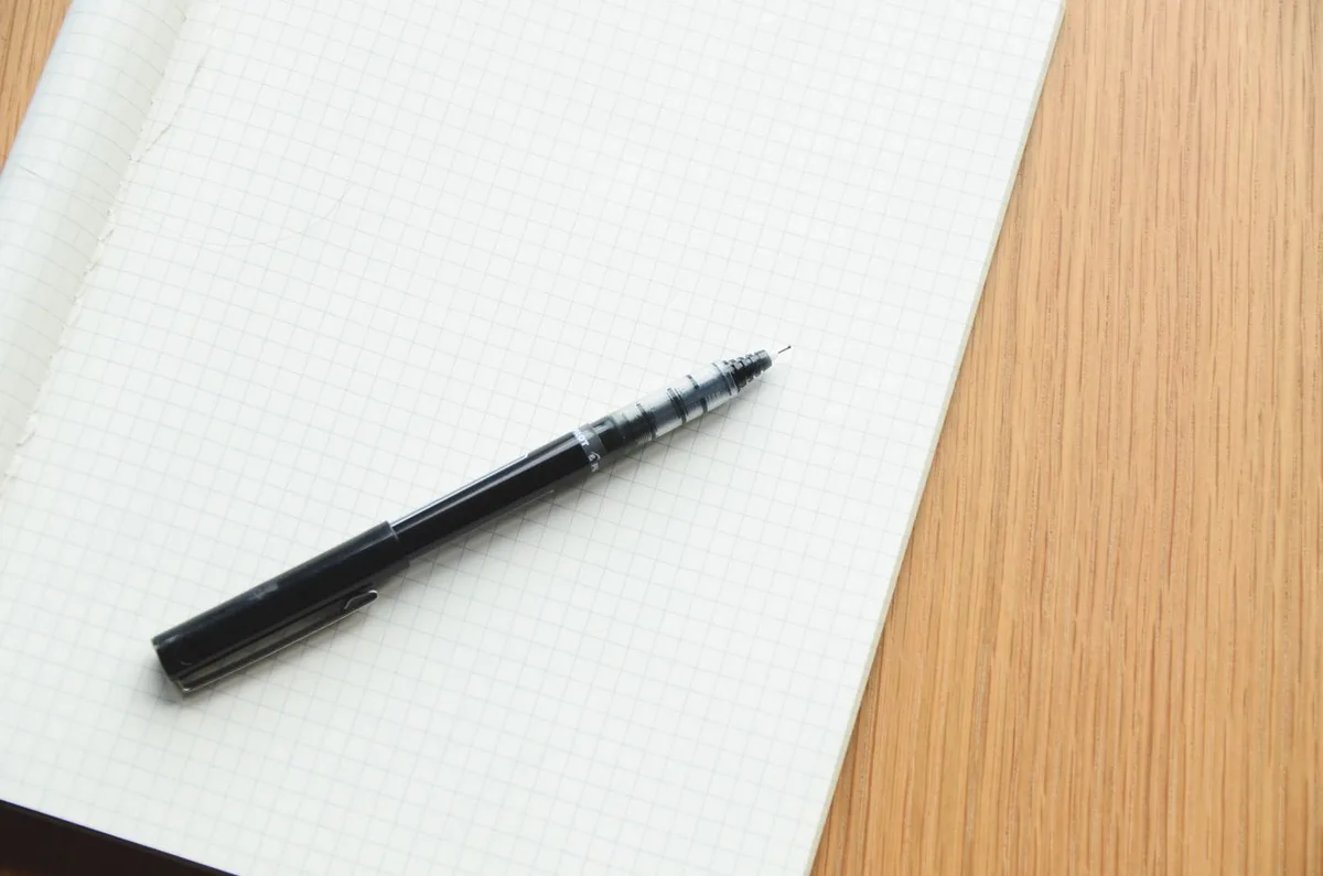 A black pen resting on an open grid-ruled notebook on a wooden desk.