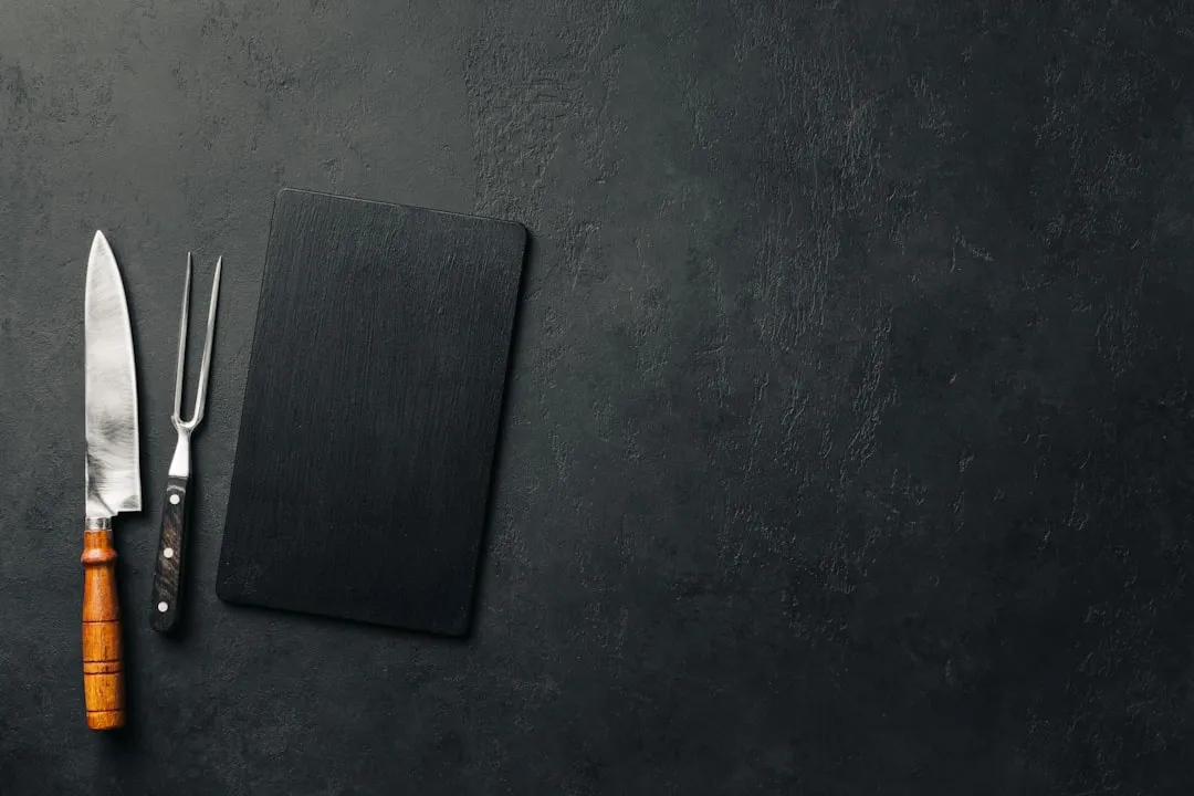 A chef's knife, a carving fork and a dark slate board laid out on a dark worktop.